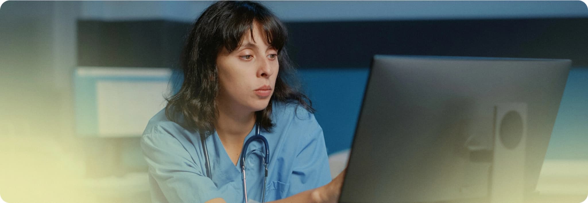 A healthcare professional in blue scrubs sits in front of a computer monitor.