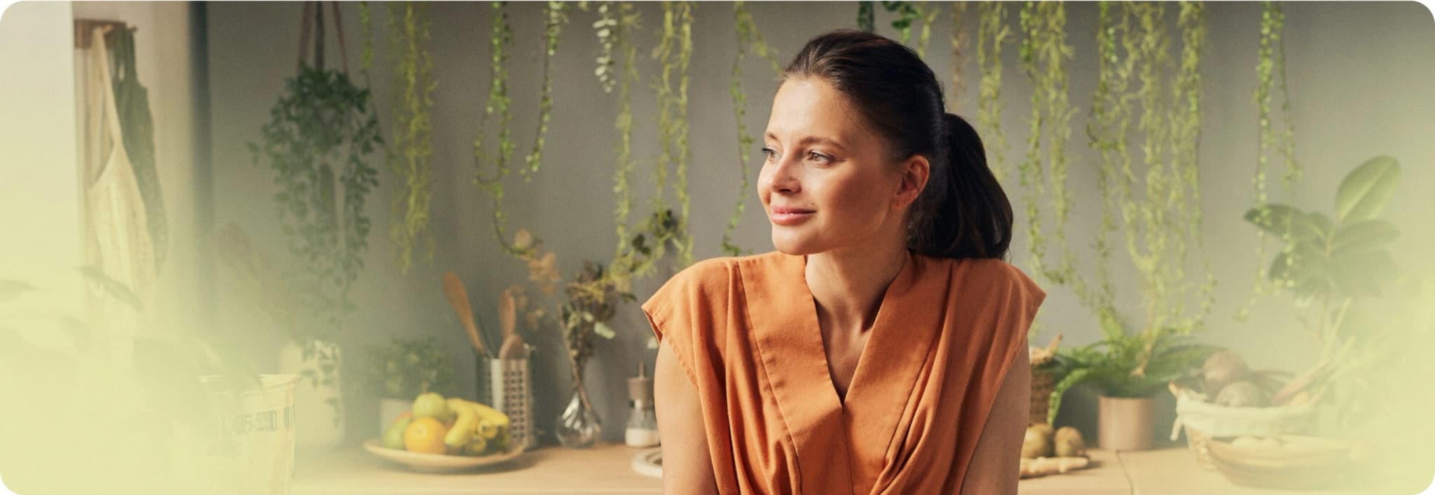 Person in orange blouse at a kitchen counter with hanging plants and fruit.